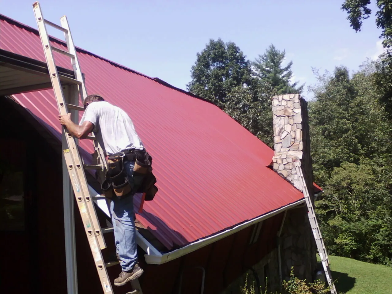 Roofer on a ladder beside a red metal roof and stone chimney — Soffit & Fascia Repair by Roofing Brothers in Newport Beach